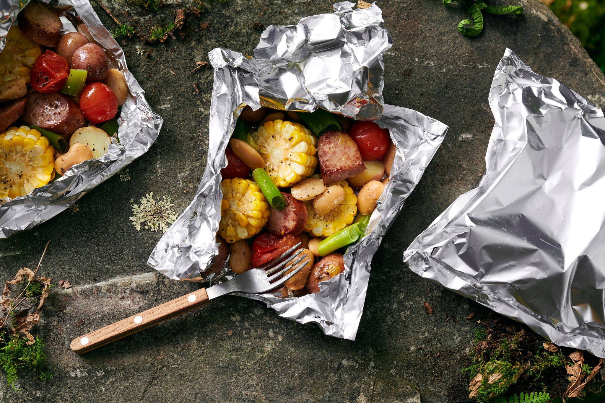 An overhead image of kielbasa and corn in a foil packet, with two other foil packets surrounding.