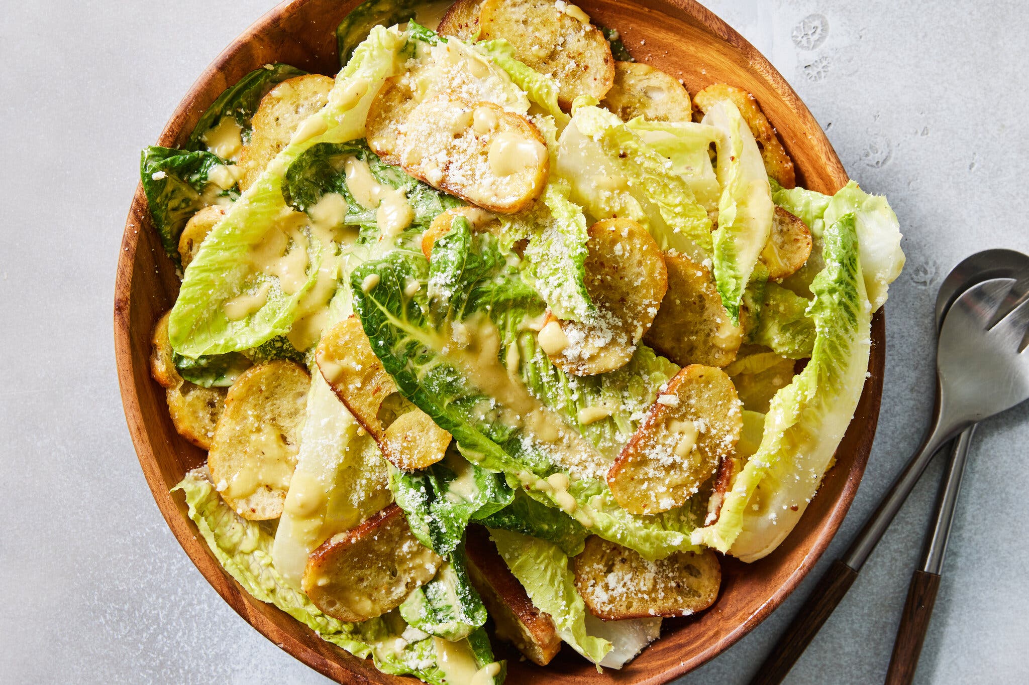 An overhead image of a Caesar salad in a wooden bowl. It’s finished with a dusting of cheese and topped with long, sliced croutons.