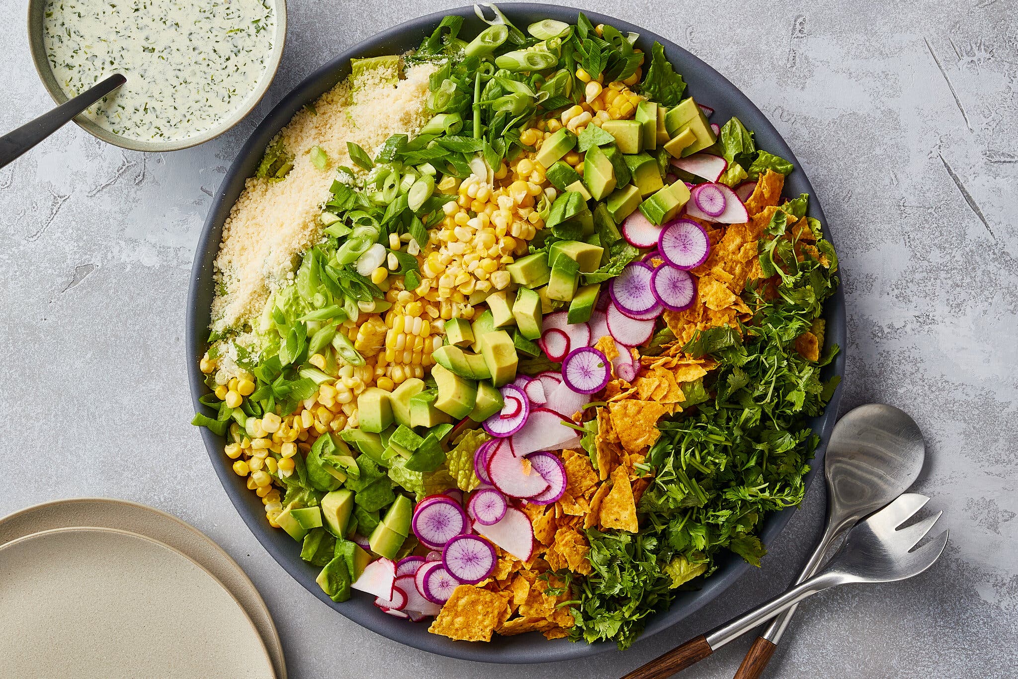 An overhead image of a salad with rows of ingredients: cheese, scallions, corn, avocado, radishes, chips and greens.