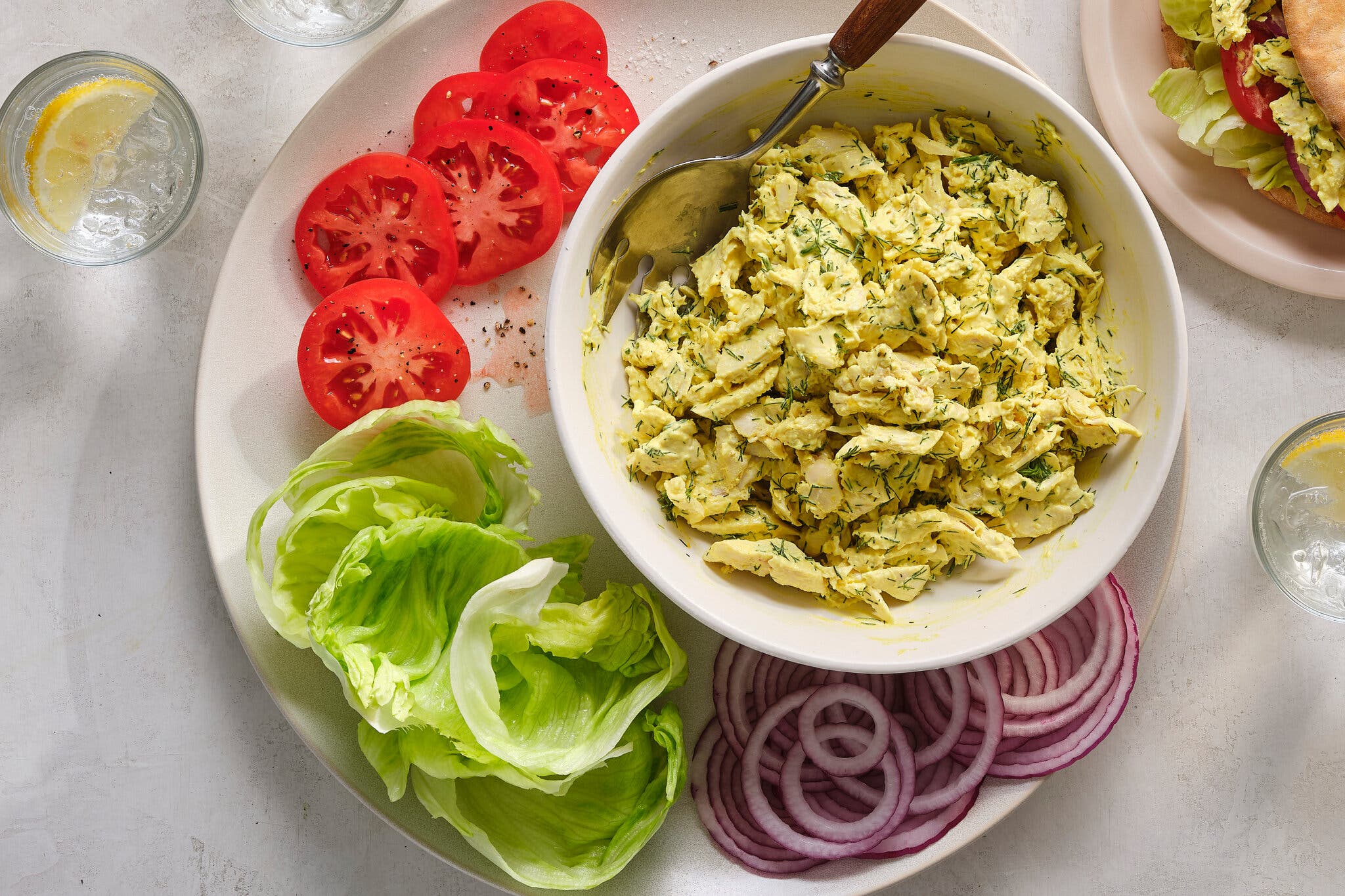 An overhead image of a bowl of chicken salad next to sliced tomatoes and onions and lettuce leaves.