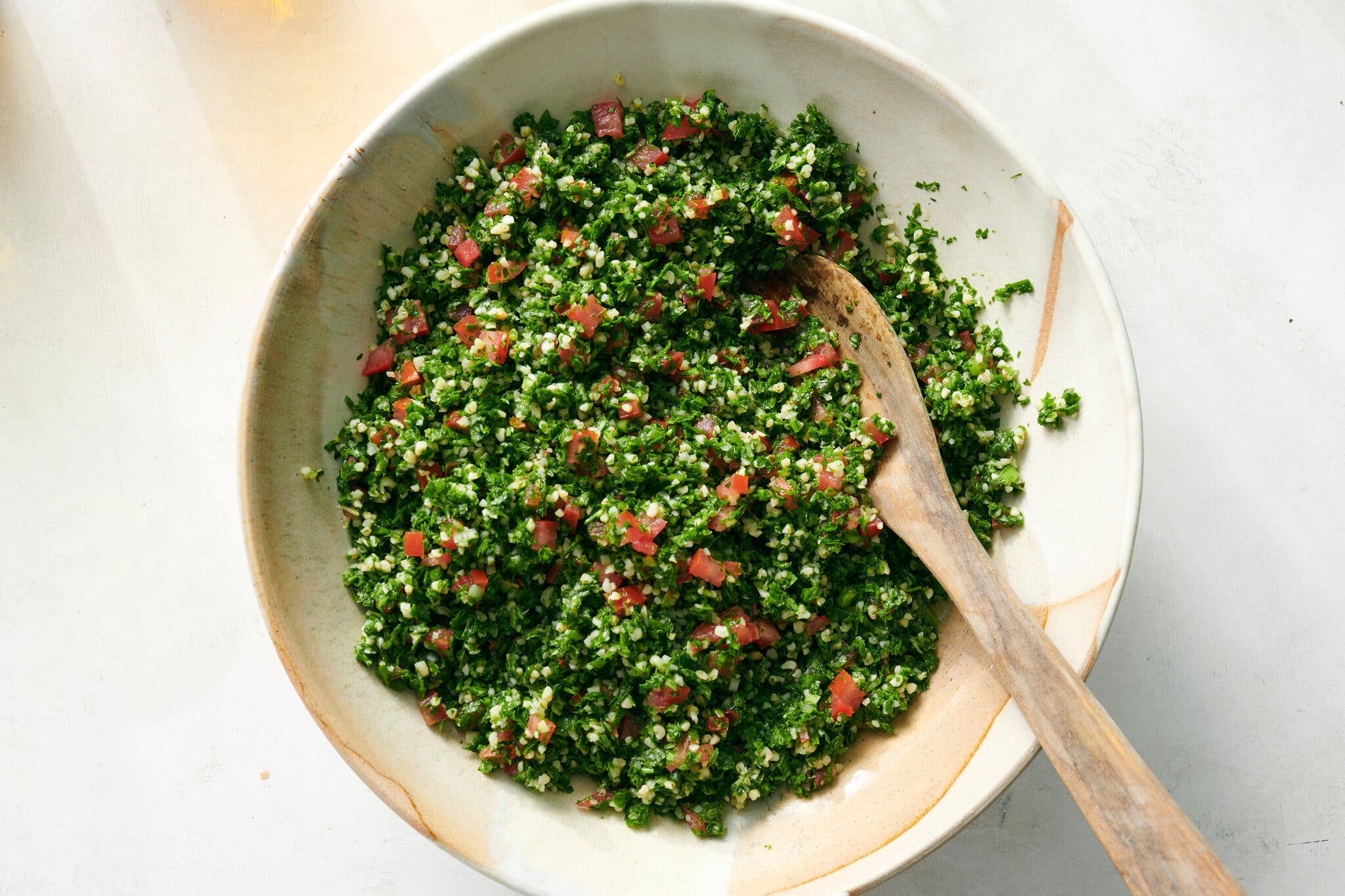 An overhead image of herby tabbouleh in a white bowl with tan accents. Tiny cubed tomatoes poke out from the bulgur-parsley mix.