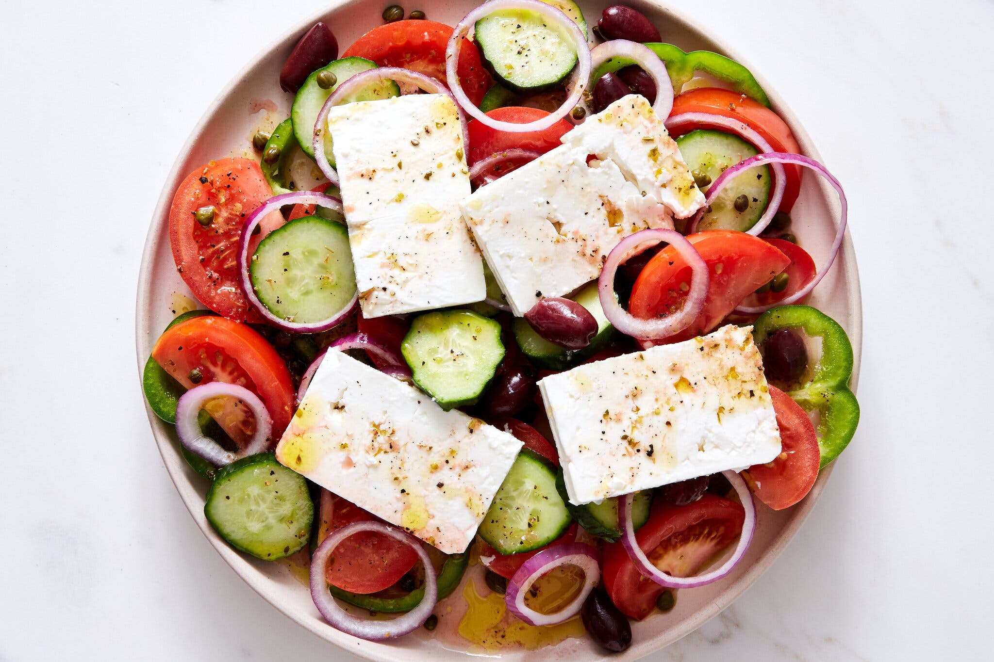 An overhead image of a pink platter topped with tomatoes, sliced cucumbers and slabs of feta.