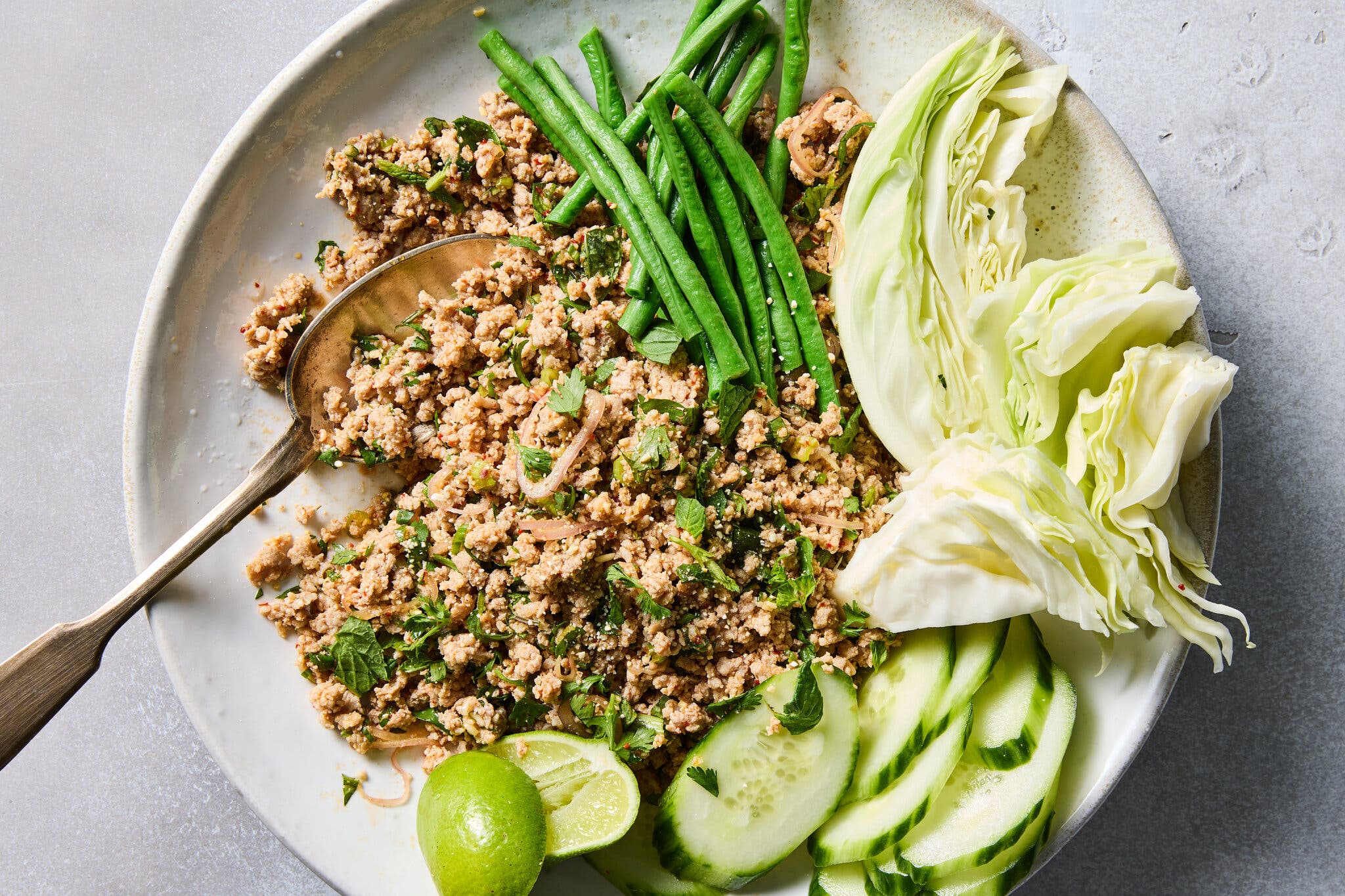 An overhead image of a platter topped with herby ground pork, sliced cucumbers, green beans and lettuce.