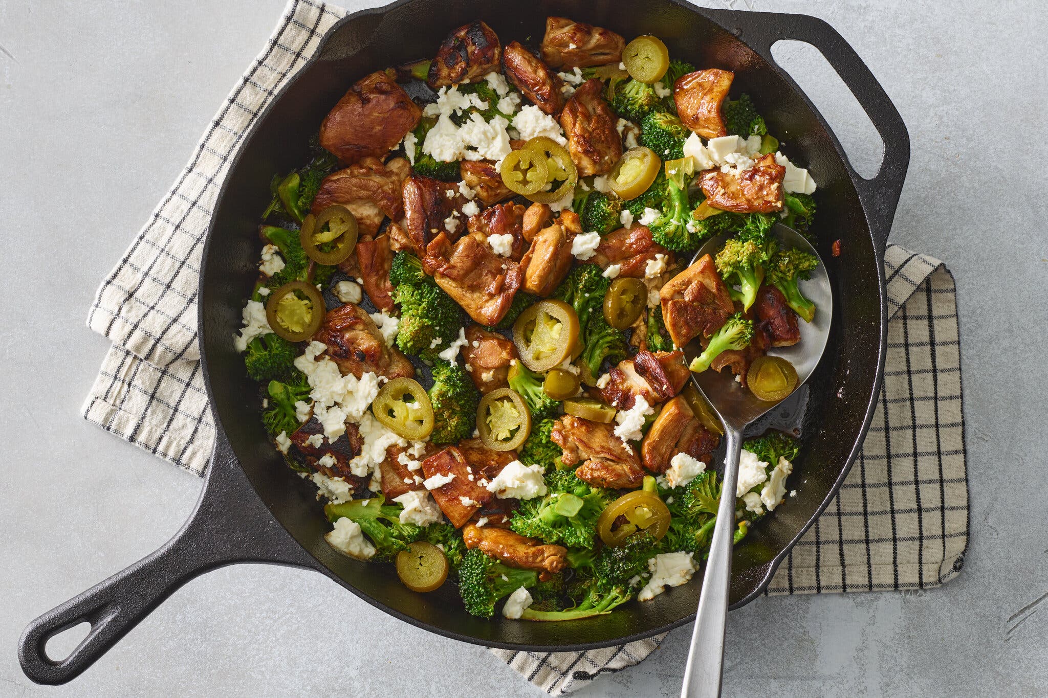 An overhead shot of a cast-iron pan on a dish towel. In the pan: sliced chicken thigh, sliced pickled jalapenos, feta cheese and broccoli.
