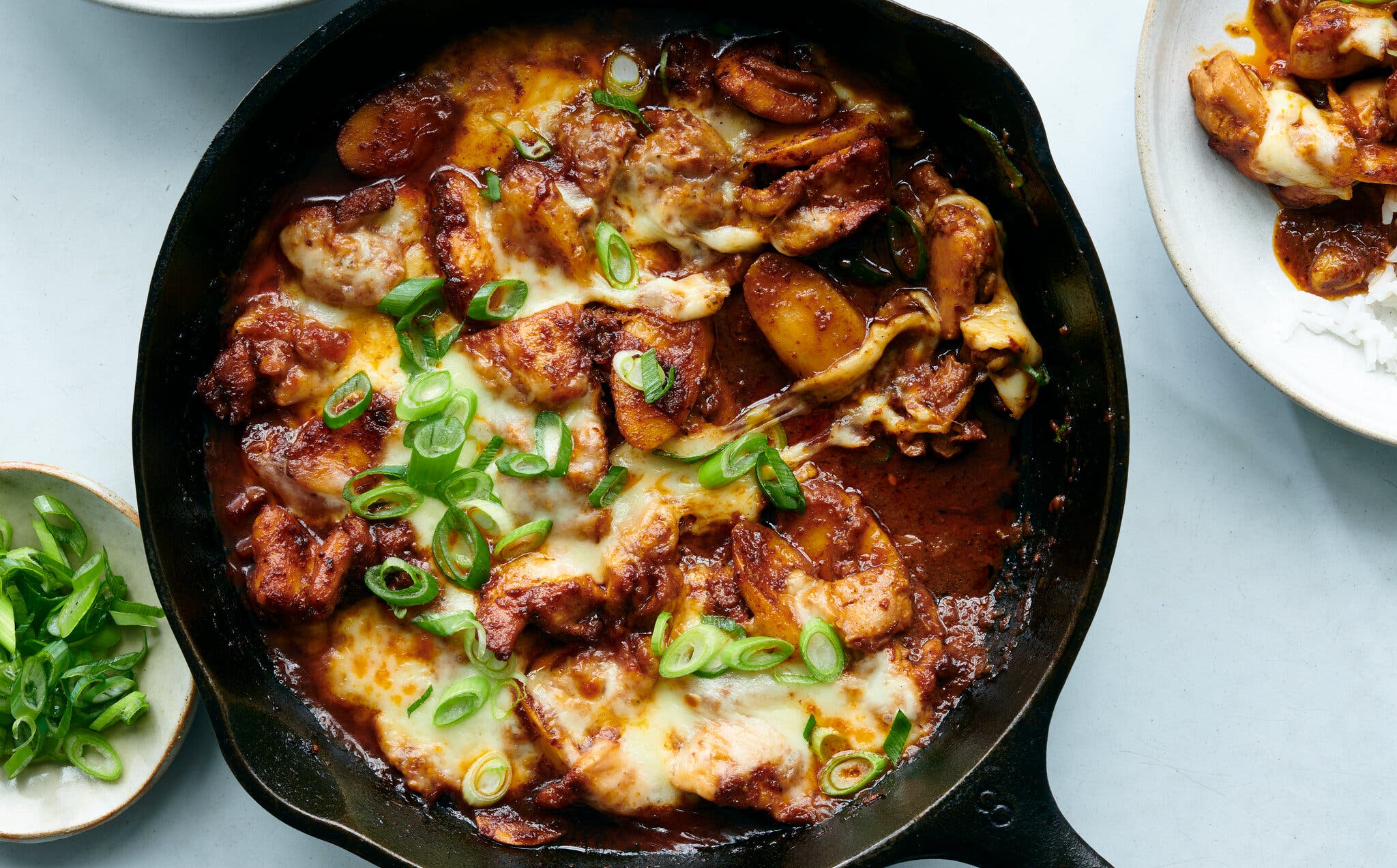 An overhead shot of a cast-iron pan with several small bowls surrounding. The pan has sliced chicken thighs covered in a sticky red sauce, garnished with sliced scallions and blanketed in melted cheese. A small bowl contains more sliced scallions, one contains rice and one has some of the chicken with rice.