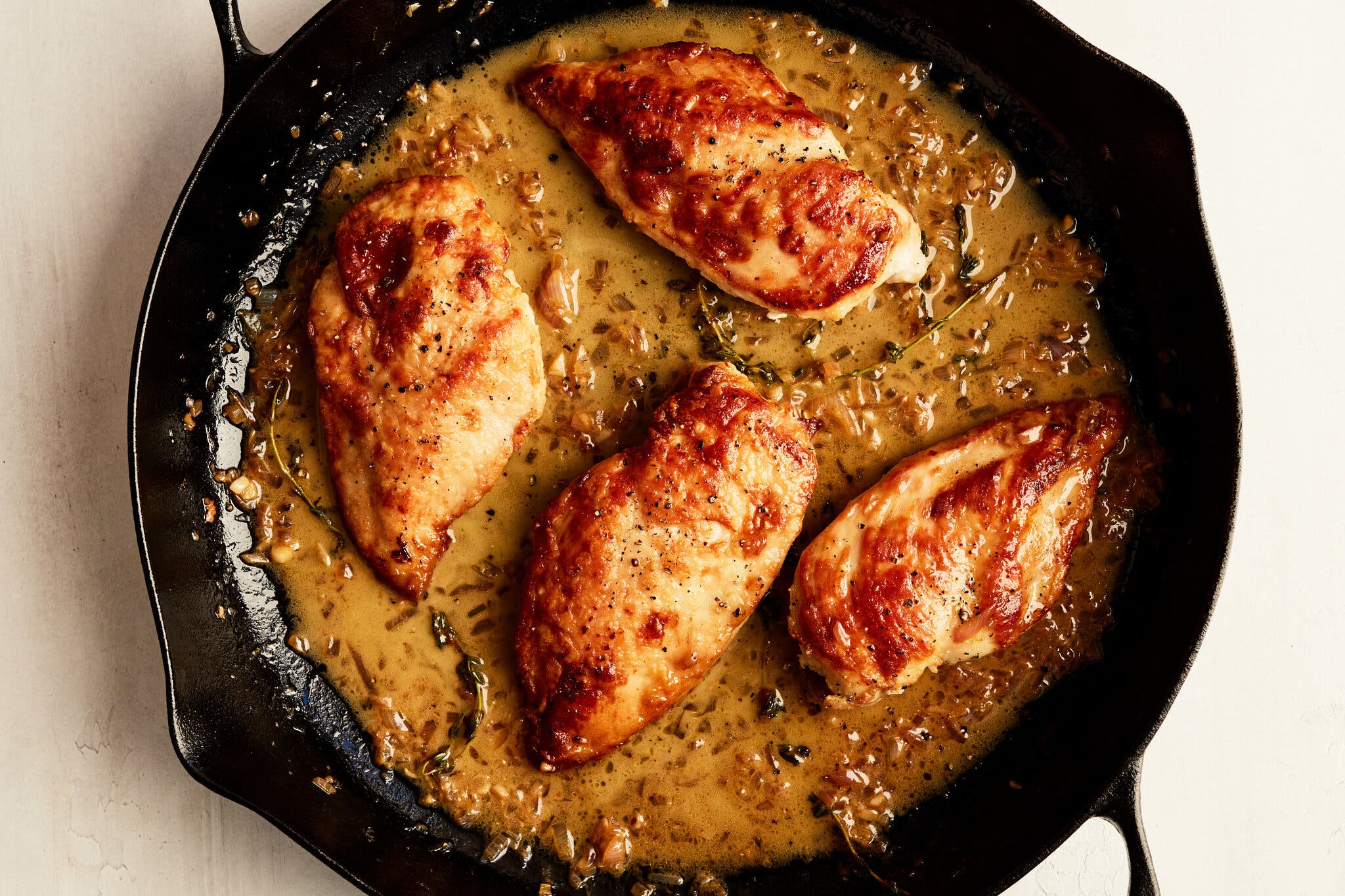 An overhead shot of a cast-iron pan with four chicken breasts, browned and covered in a lemony sauce with thyme sprigs.