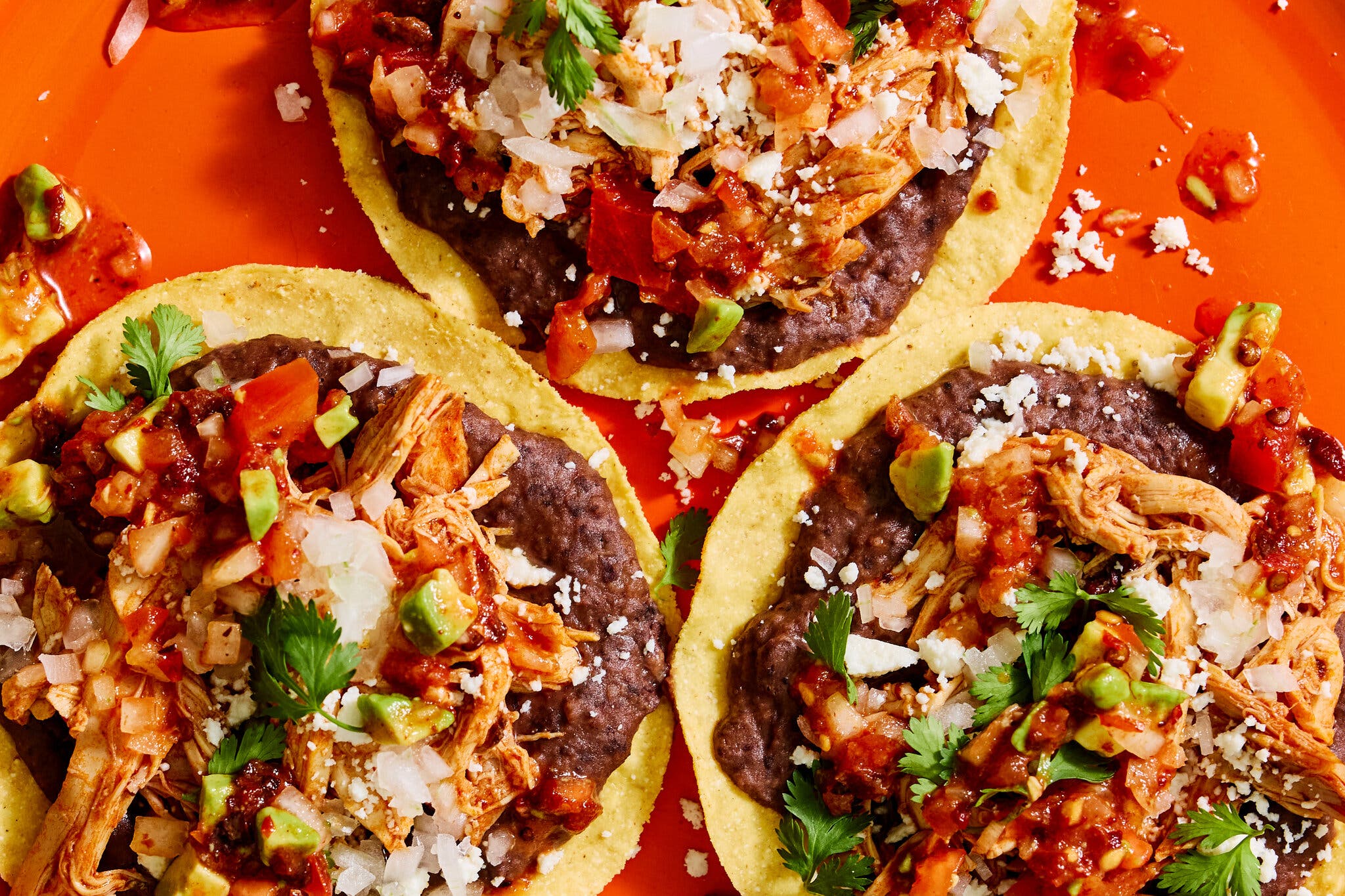 An overhead shot of three tostadas, layered with refried beans, chicken tinga, cilantro, avocado and diced white onion,