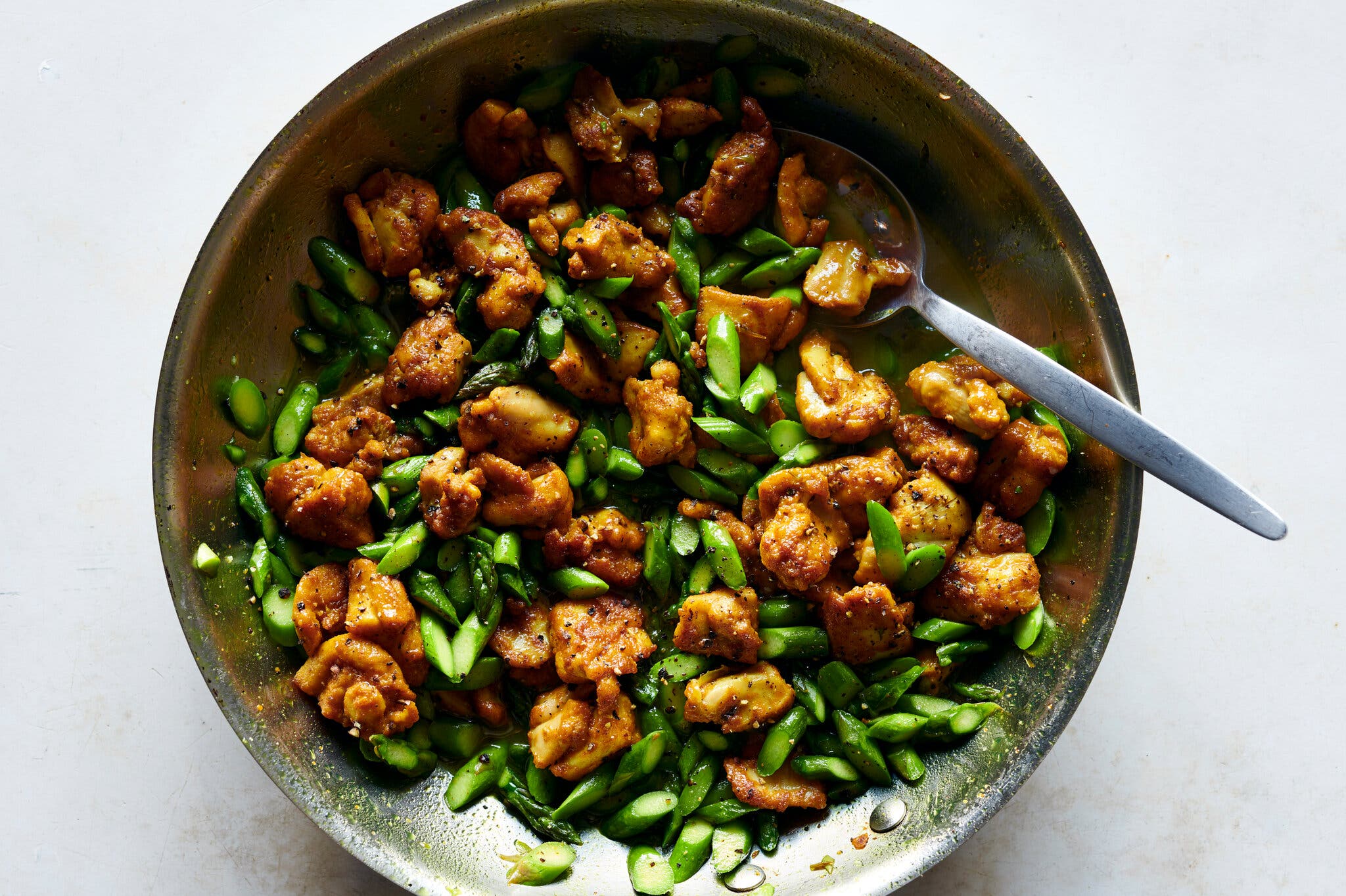 An overhead shot of a pan with sliced chicken thighs and sliced asparagus. There is a spoon in the pan.