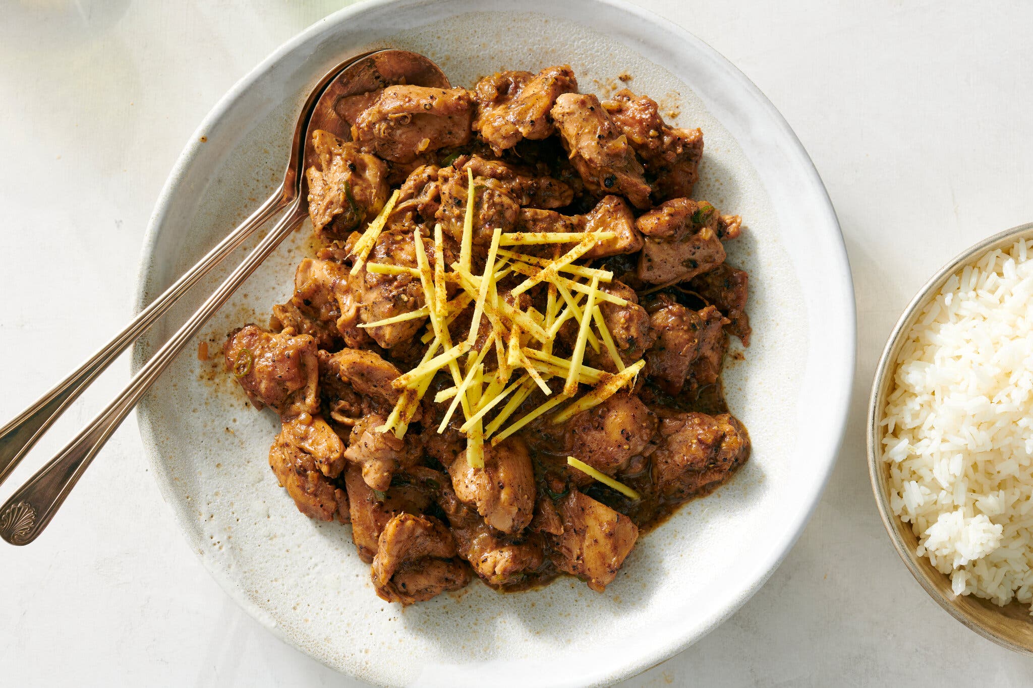 An overhead shot of a serving plate with browned sliced chicken thighs, garnished with black pepper and sliced ginger on top. There are two serving spoons in the plate, and a small bowl of rice to the side.