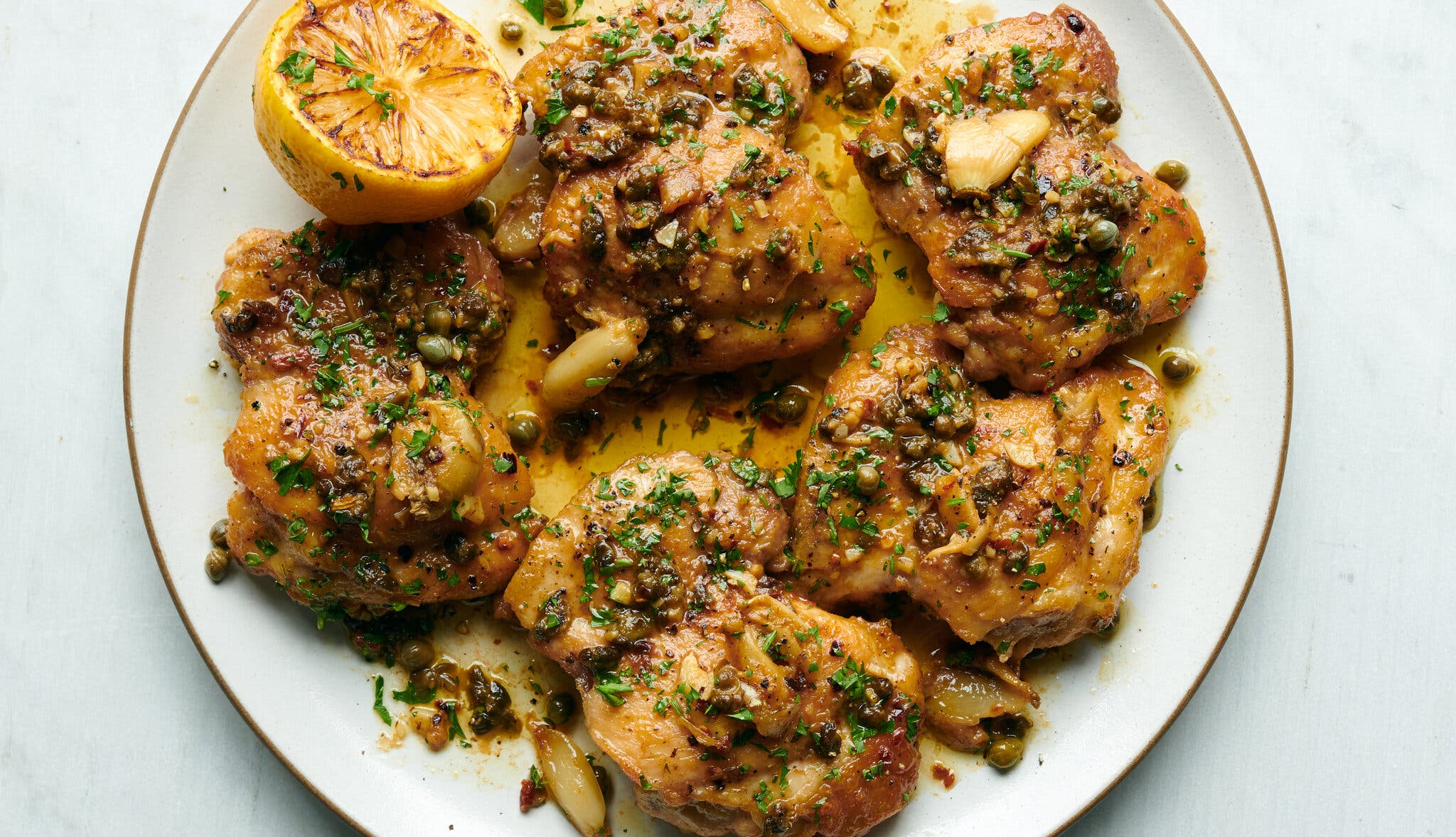An overhead shot of a serving plate with browned chicken thighs covered in a lemony sauce with parsley, capers and garlic. There is a lemon half on the plate.