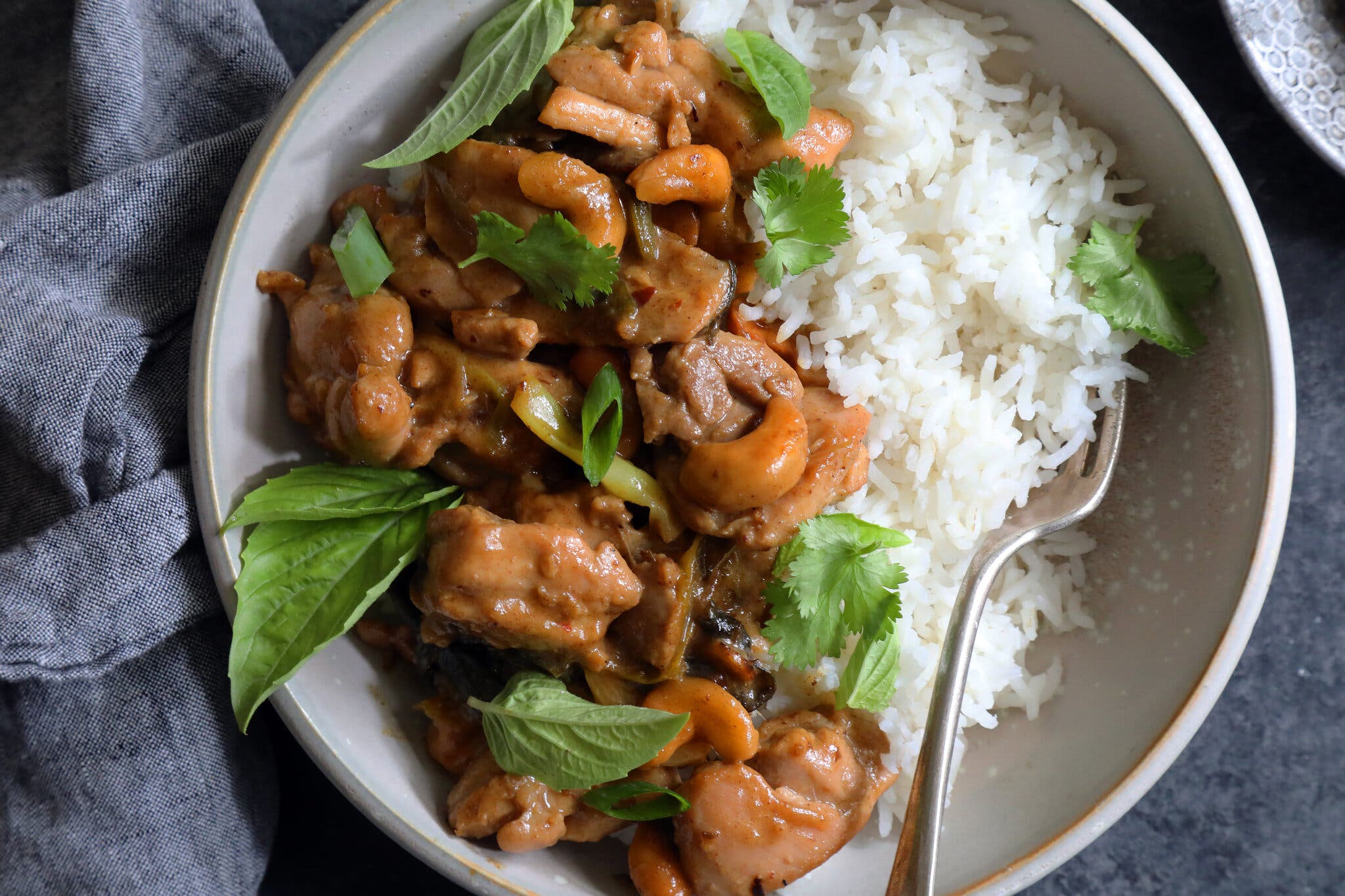 An overhead shot of a bowl with rice with sliced chicken thighs, with cashews and dates, garnished with mint and cilantro. There is a fork in the bowl.