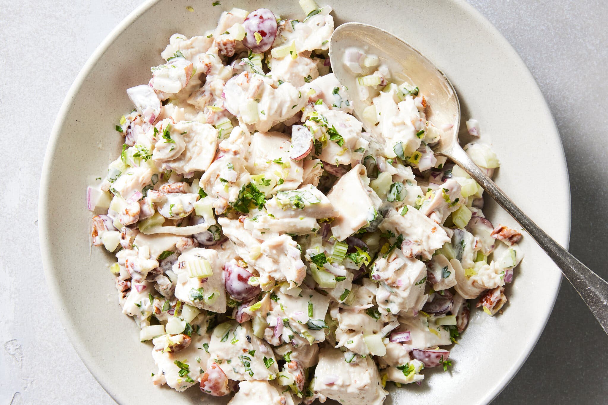An overhead shot of a bowl with chicken salad with diced chicken, grapes, celery and red onion. There is a spoon in the bowl.
