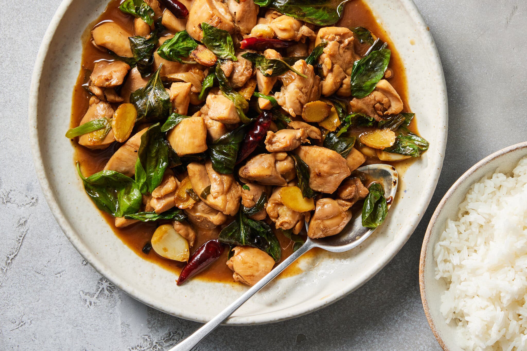 An overhead shot of a serving plate with sliced chicken thighs in a brown sauce, with leaves of basil, ginger coins and red chiles. There is a spoon in the plate and a bowl of rice to the side.