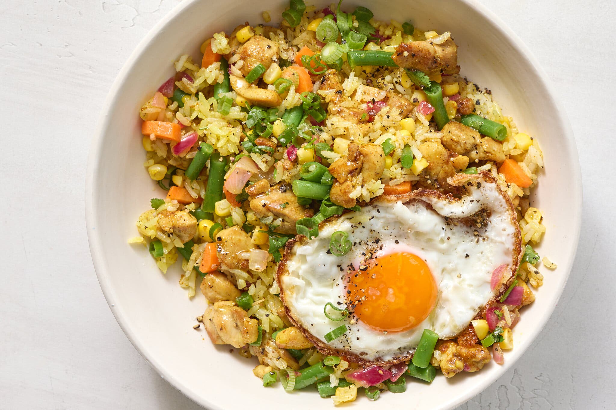 An overhead shot of a bowl with yellow-tinted fried rice with pieces of chicken, green beans carrots, corn, scallions, topped with a fried egg.