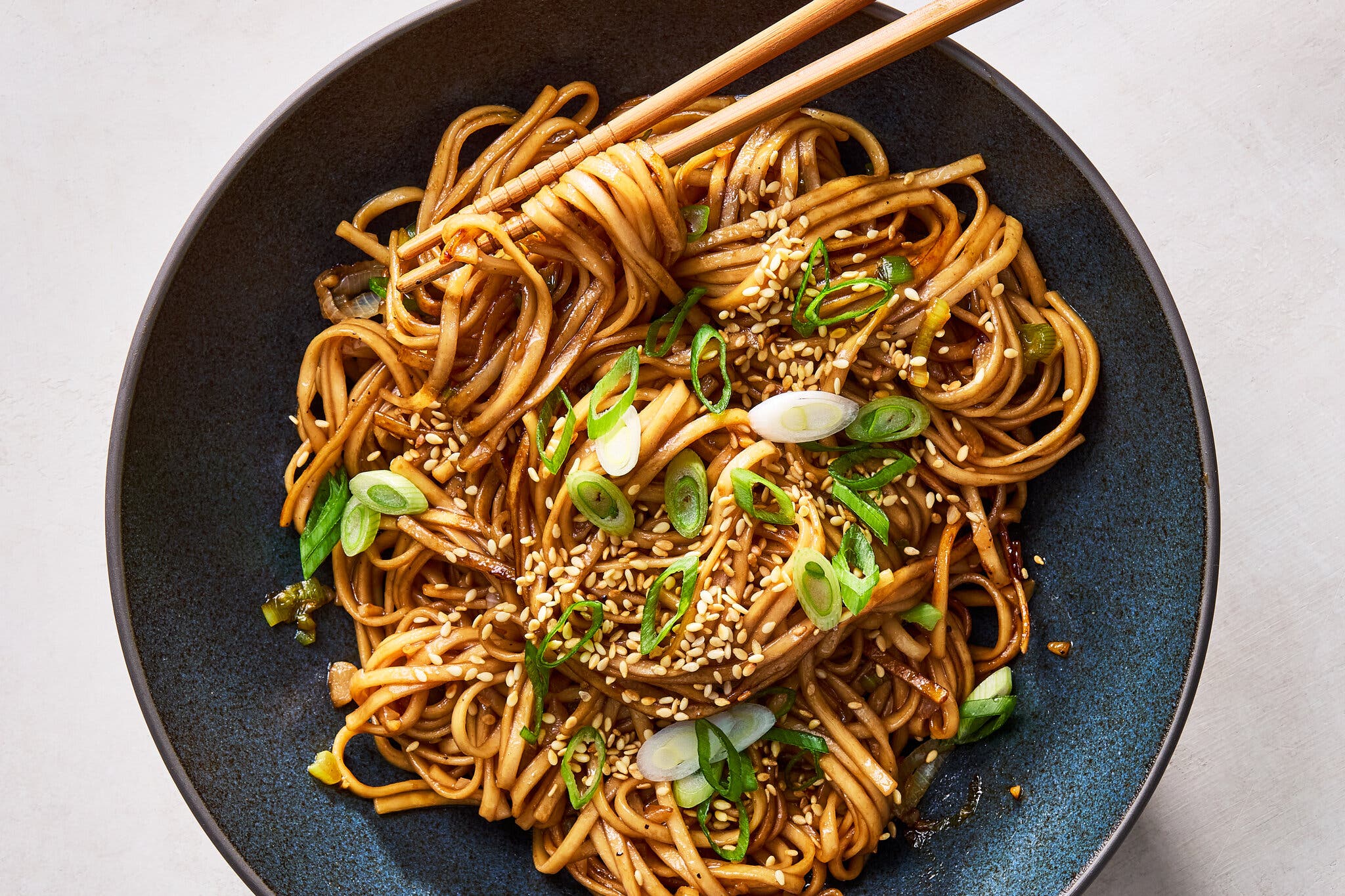 An overhead image of a dark blue bowl filled with sauce-slicked noodles topped with sesame seeds and slivered scallions.