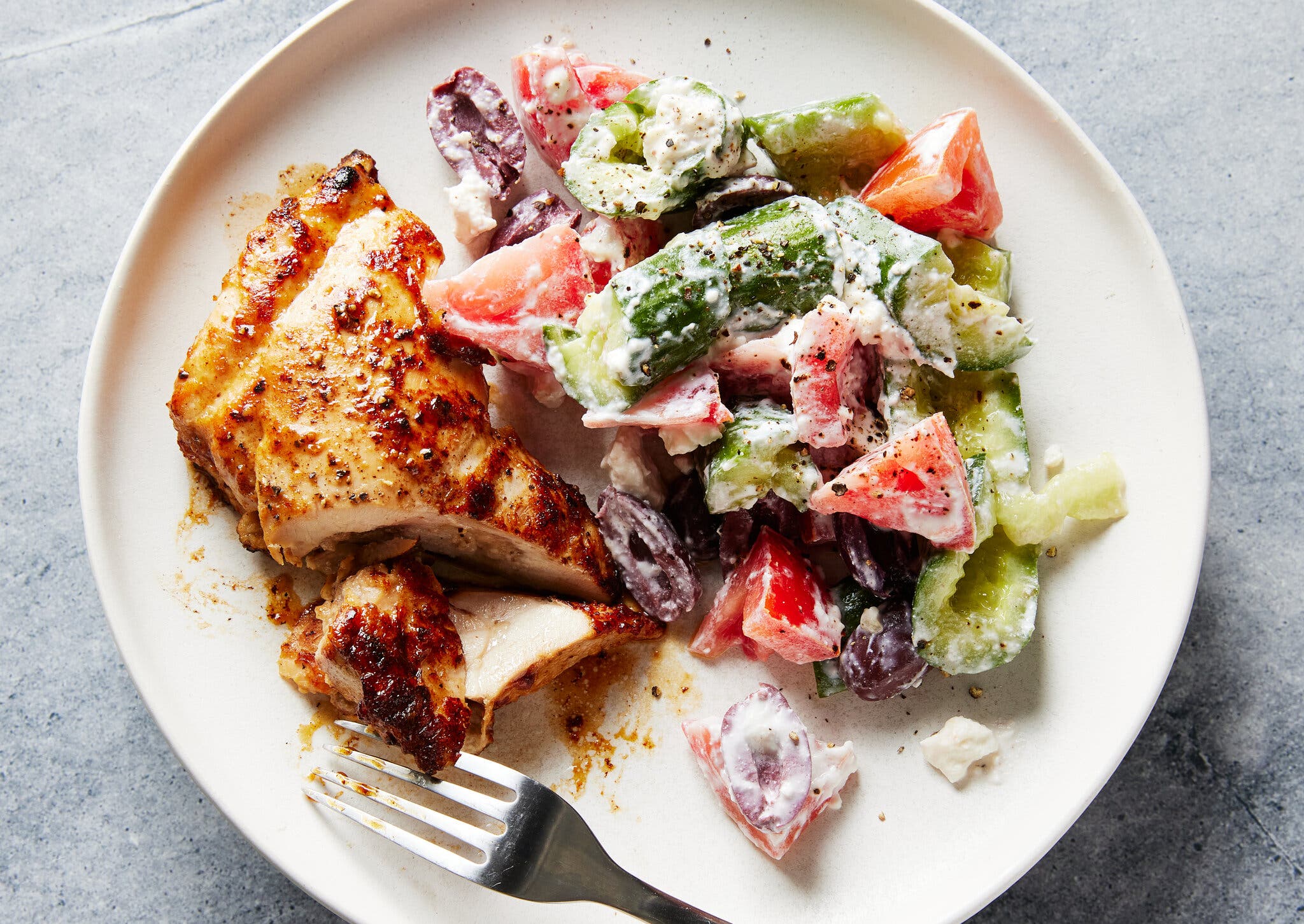 An overhead shot of a plate with sliced, spiced chicken thigh, a fork and a side salad of cucumbers, tomatoes and olives covered in a feta dressing.