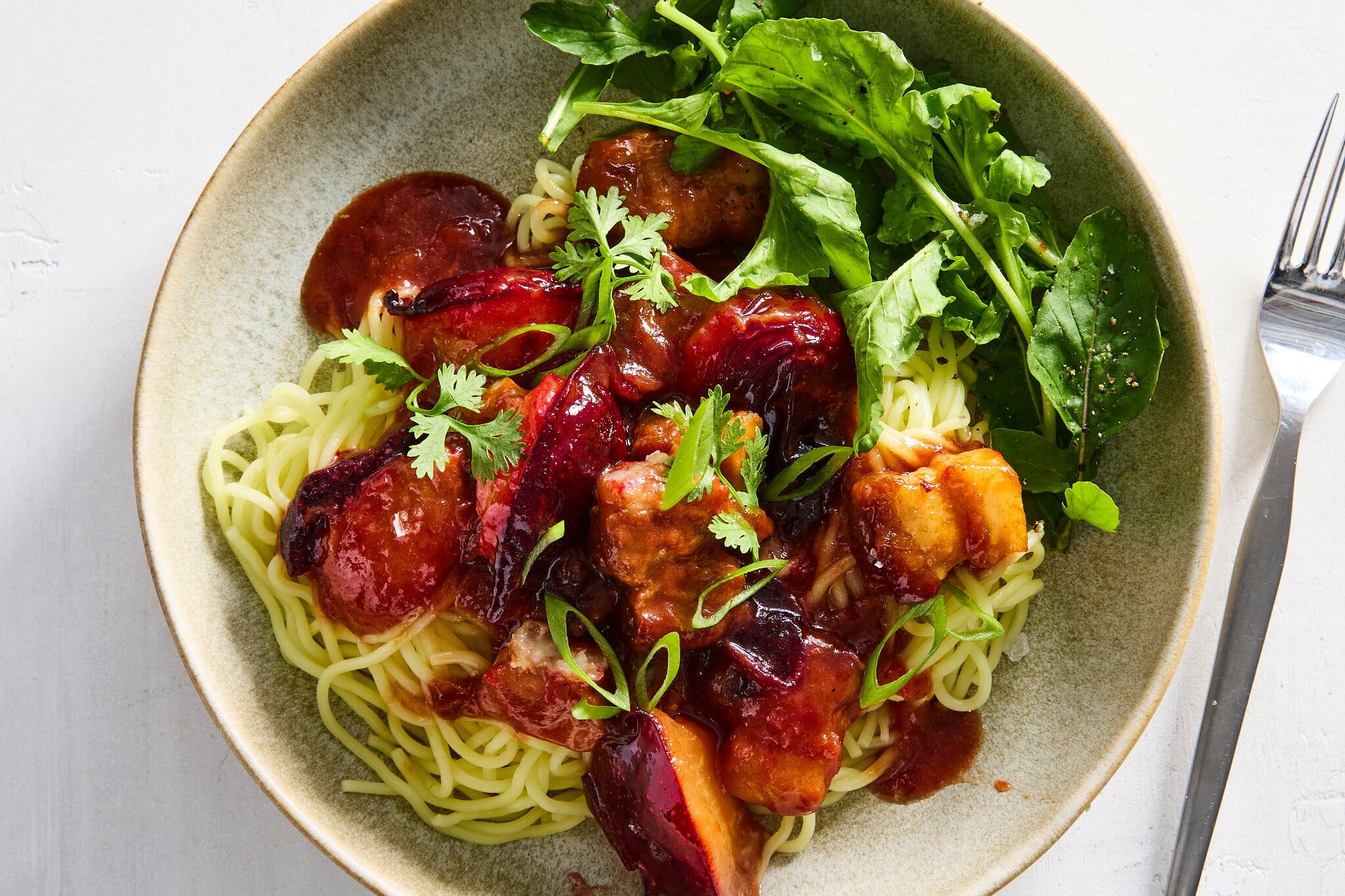 A bowl with stir-fried pork belly and plums over noodles with greens alongside.