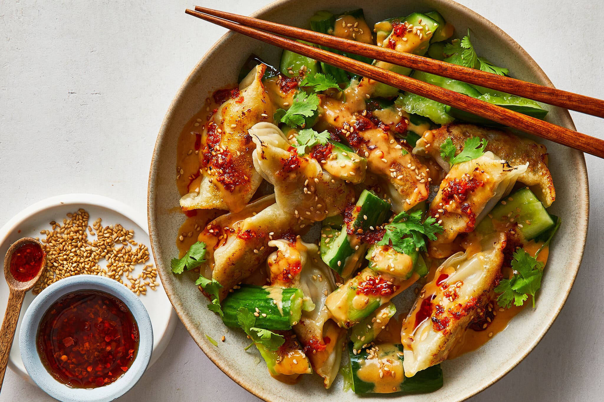 An overhead image of a beige bowl filled with dumplings, cucumber chunks and cilantro. Everything is drizzled with a light brown peanut sauce, a red sauce with chile flakes and sprinkled with sesame seeds.