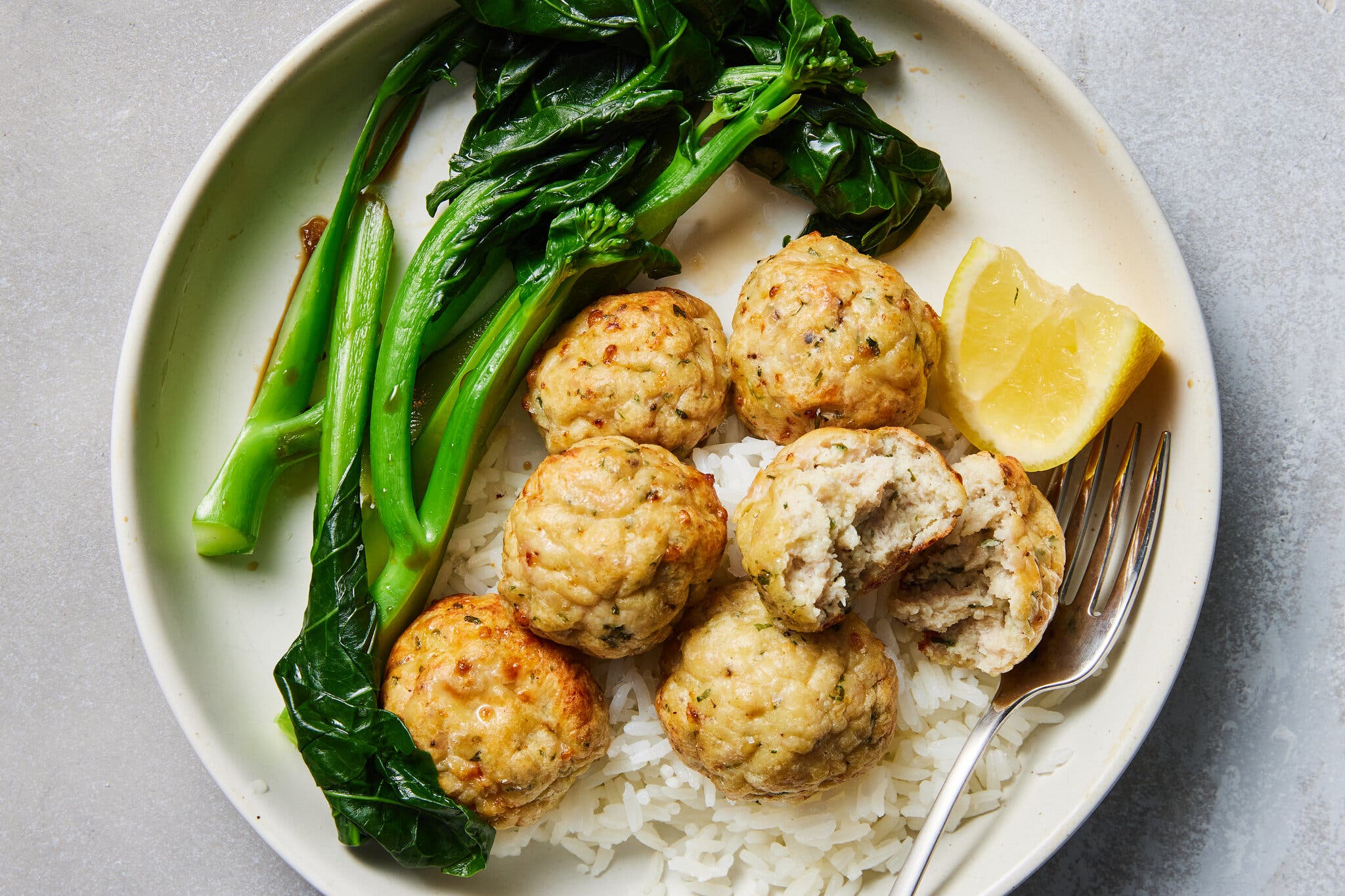 An overhead image of a white plate filled with brown meatballs atop white rice, with a side of green broccolini and a squeezed lemon wedge.  