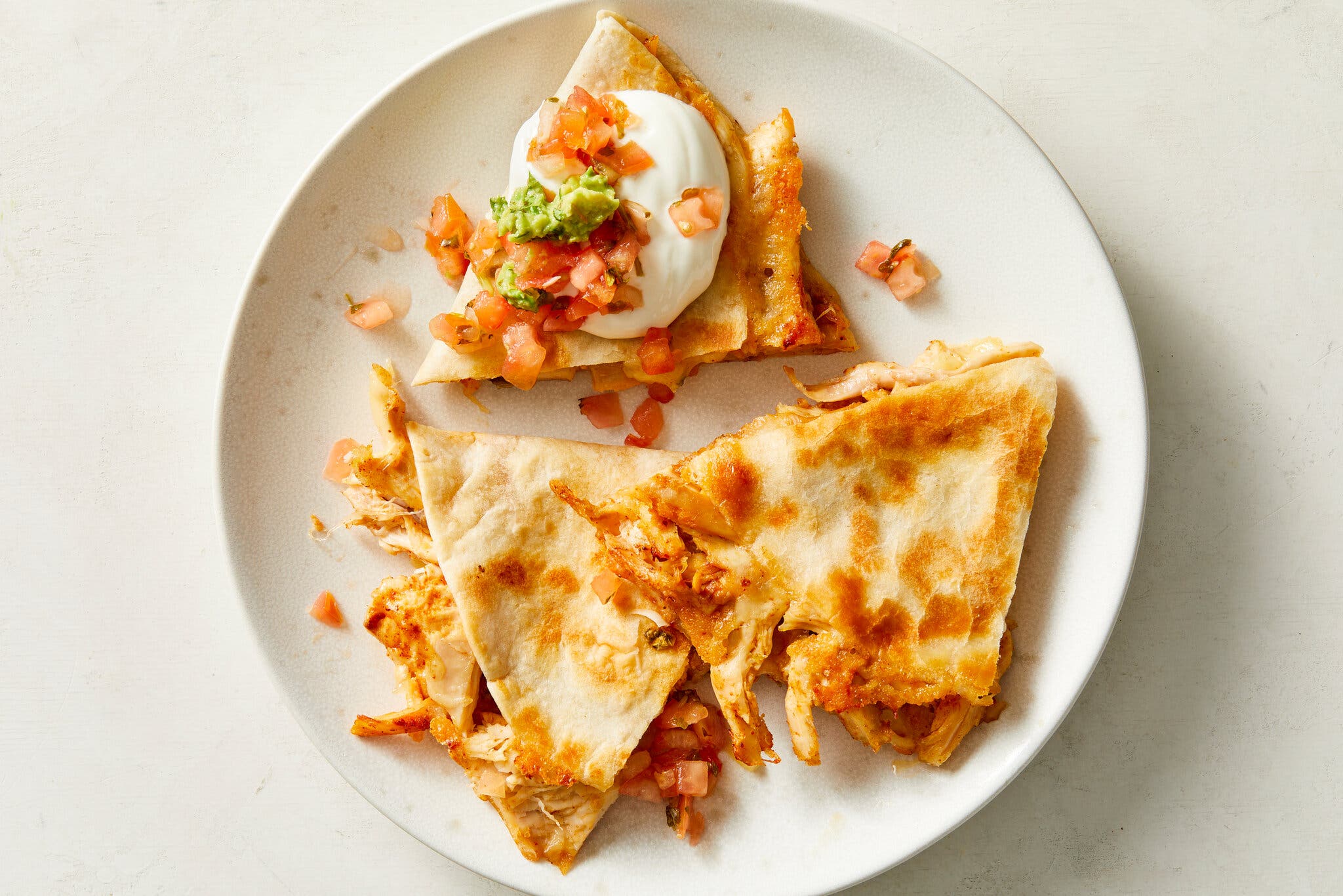 An overhead image of three tortilla triangles stuffed with shredded chicken and cheese. Sour cream, salsa and guacamole top one of the triangles.