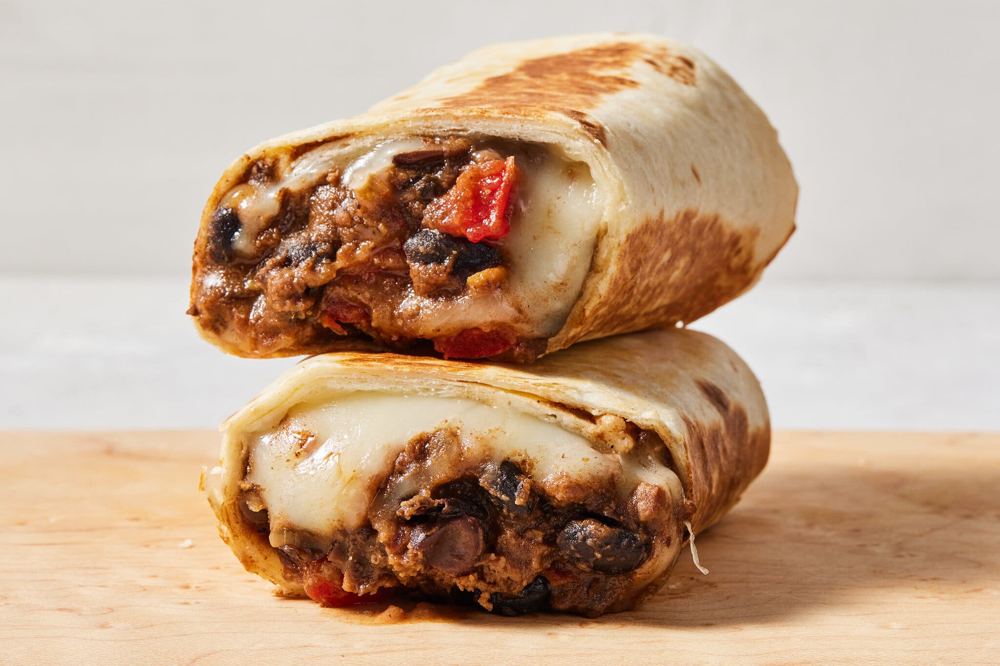 An image of two halves of a burrito stacked on top of one another so you can see the filling, which is facing the camera: a mixture of brown-red ground meat, beans and melty white cheese. 