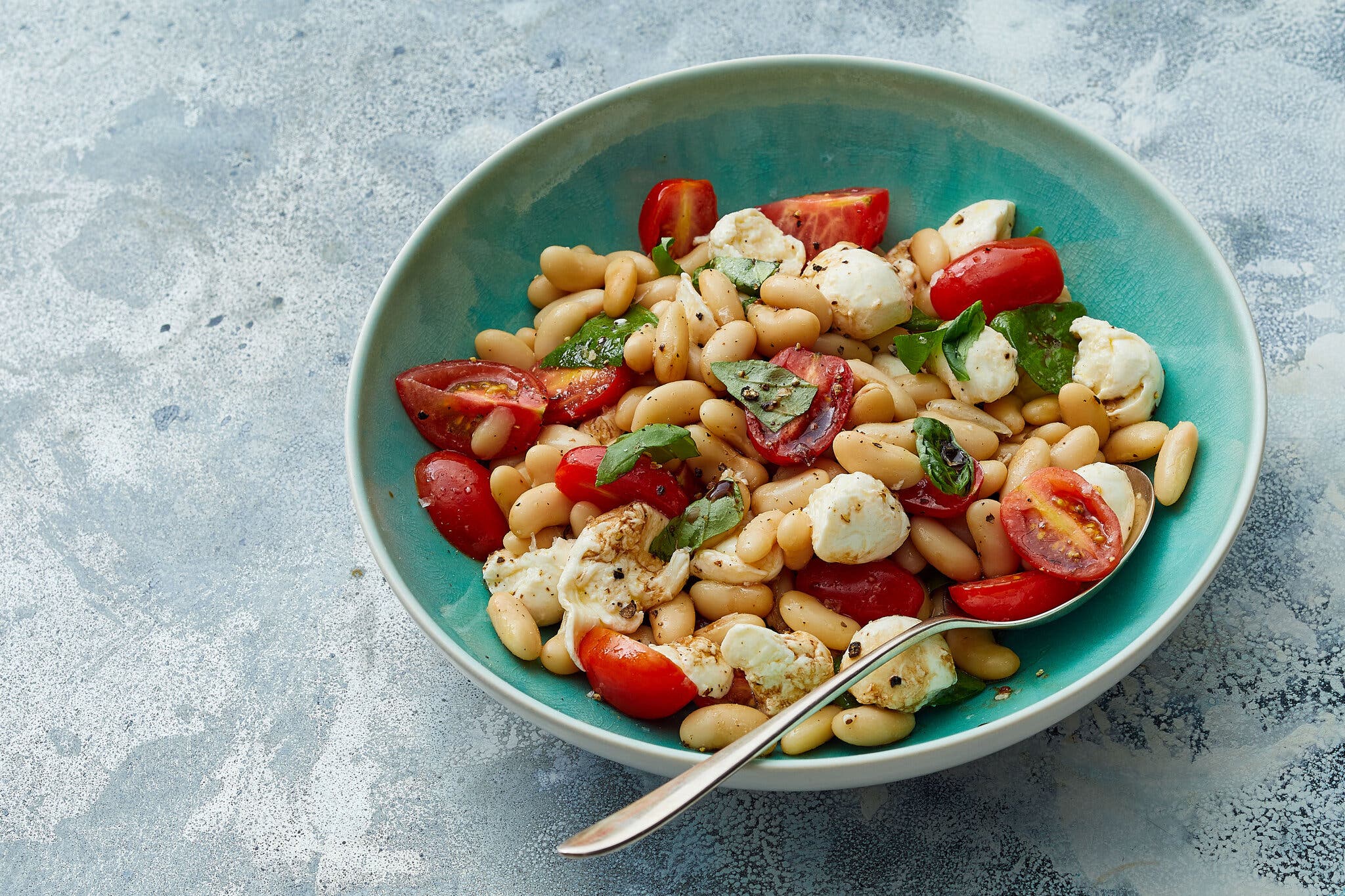 An overhead image of a teal green bowl filled with white beans, white mozzarella balls, cut red cherry tomatoes and torn green basil leaves.