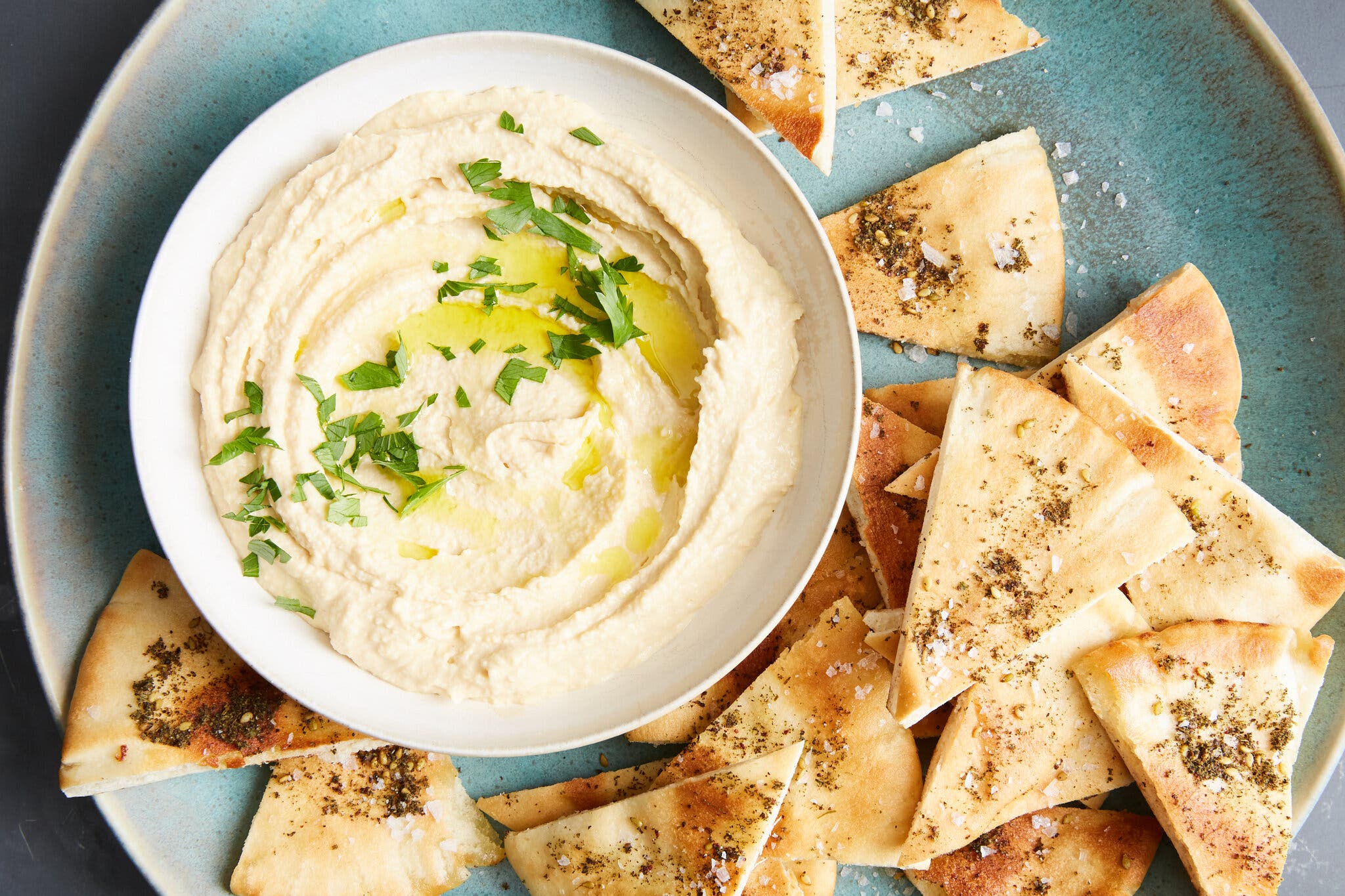 An overhead image of a white bowl filled with creamy beige hummus drizzled with olive oil and chopped green parsley. Pita wedges topped with dried herbs and flaky sea salt are on the side. 