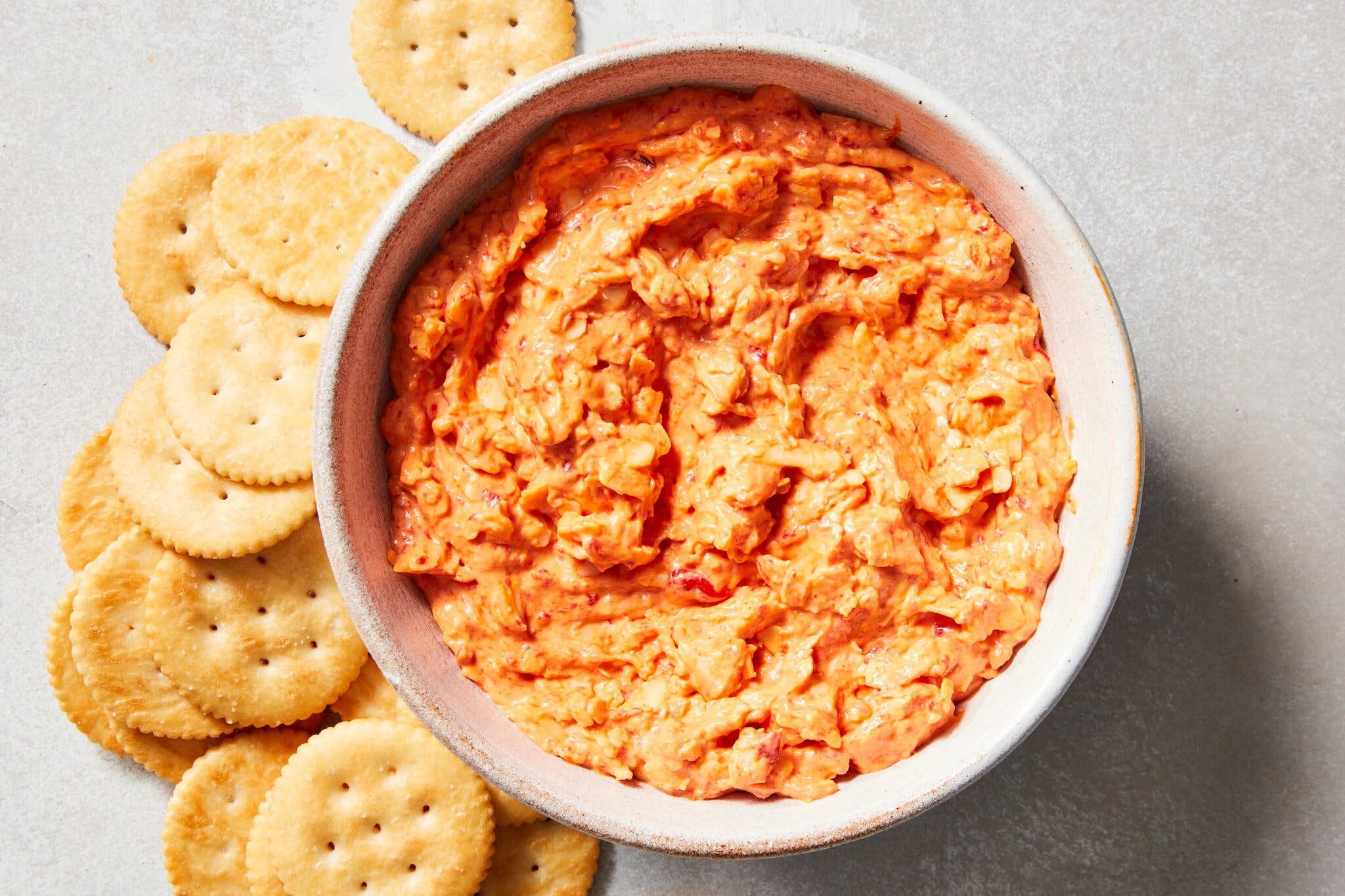 An overhead image of a white bowl filled with creamy orange pimento cheese dip, with round golden brown crackers on the side. 