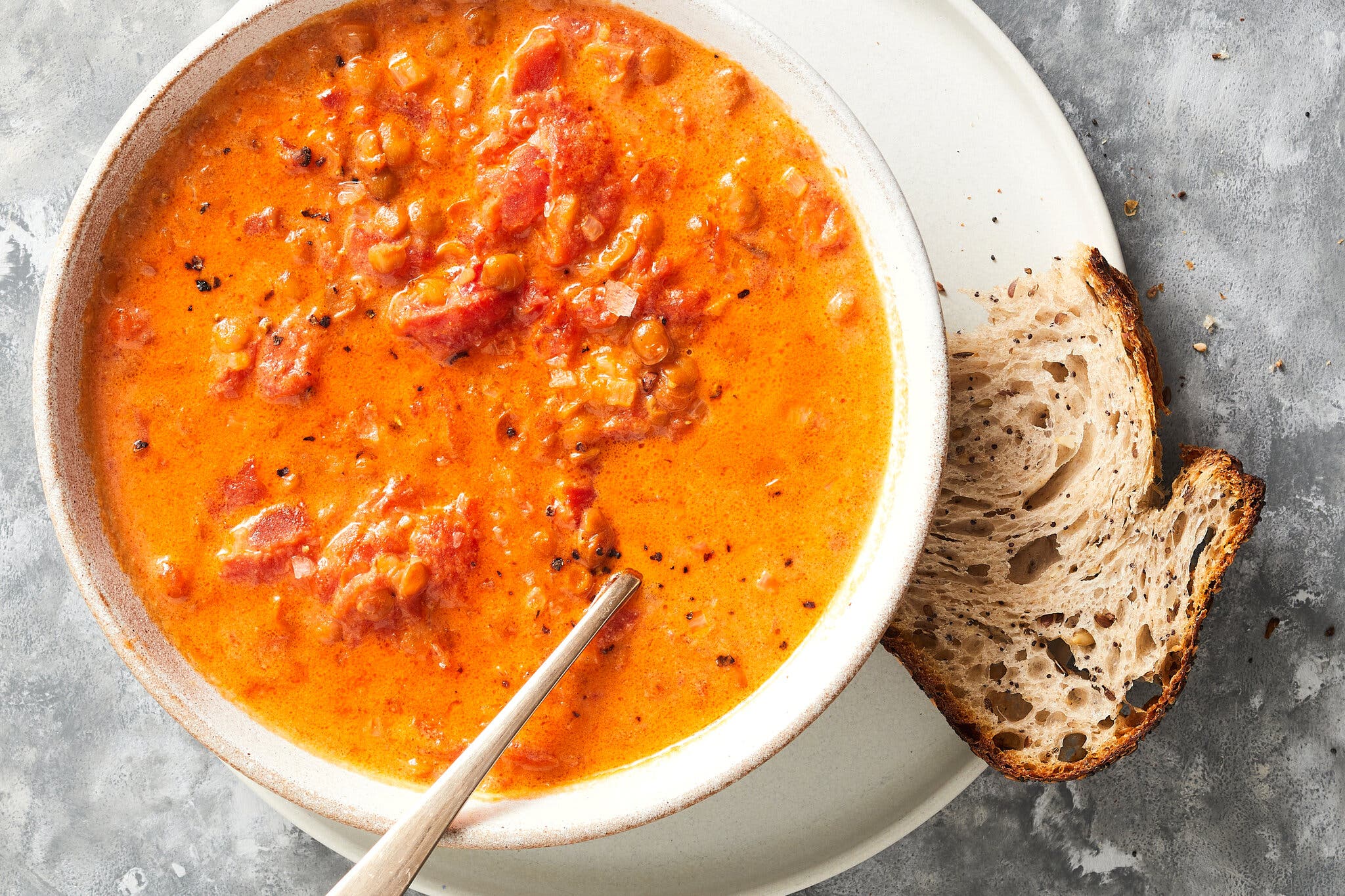 An overhead image of a white bowl filled with creamy red-orange soup with lentils and tomatoes and specks of black pepper. A slice of crusty bread is on the side. 