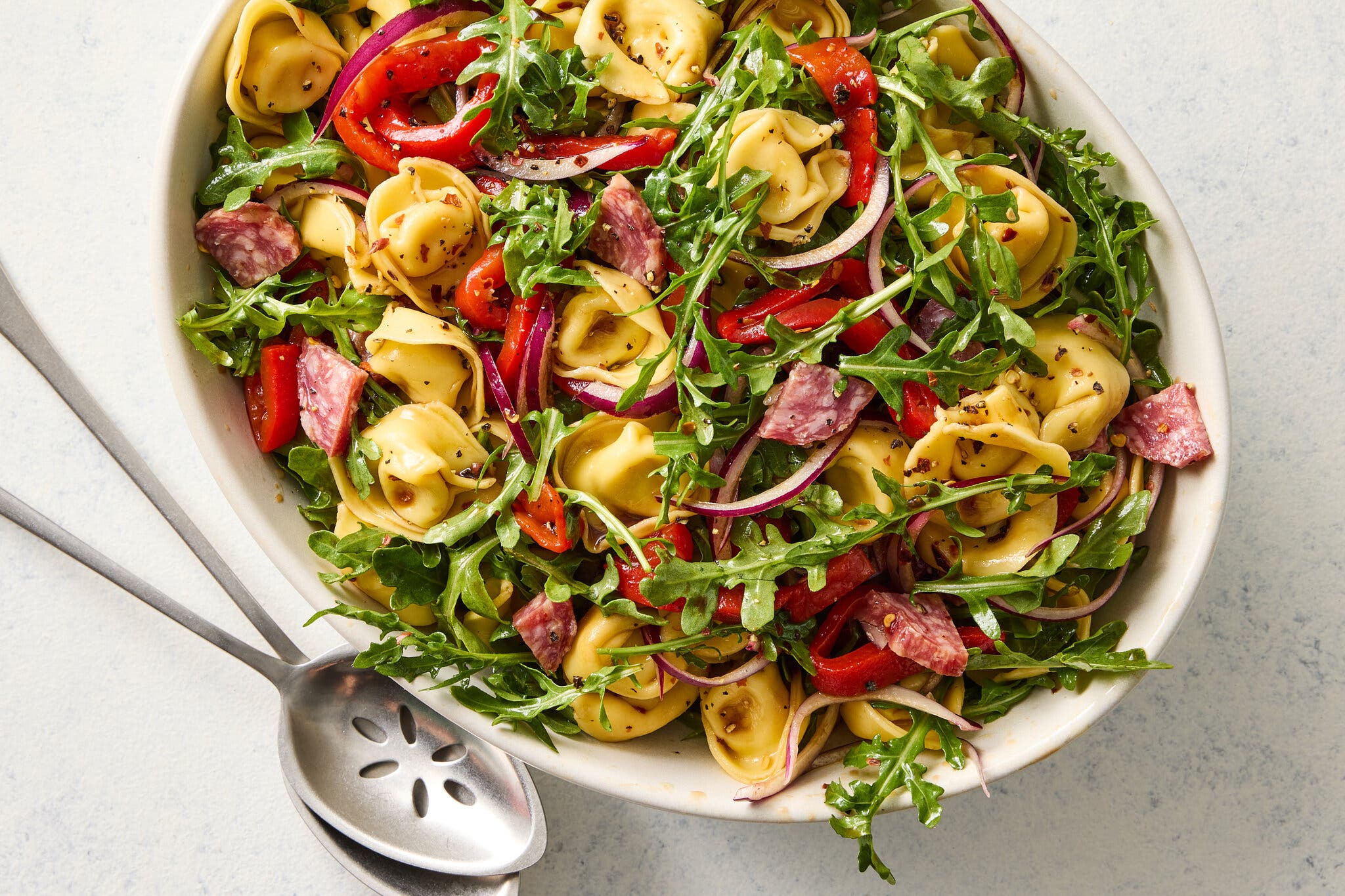 An overhead image of a white oval serving bowl filled with round tortellini pasta, red peppers, salami chunks and arugula leaves.