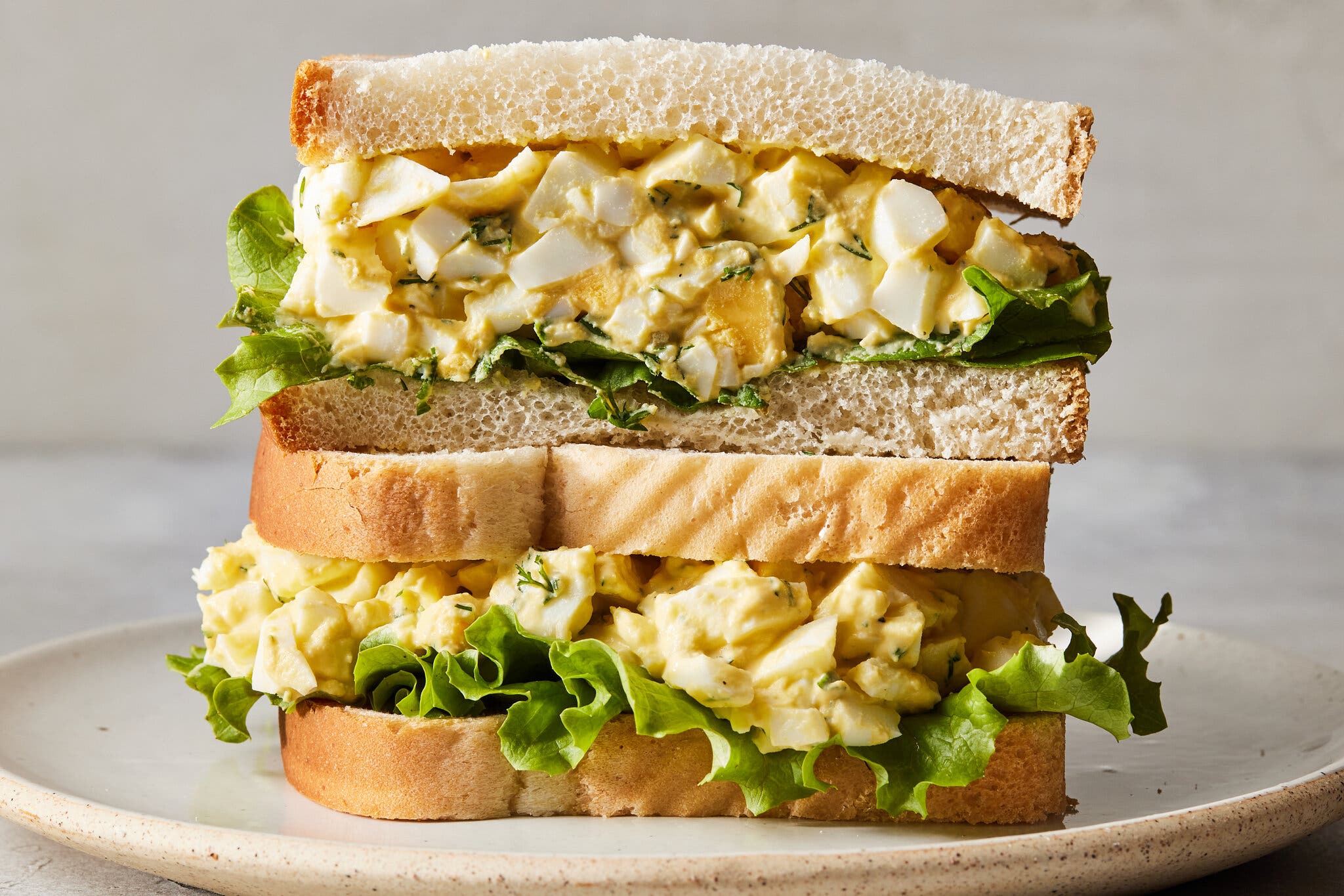 A close-up image of the side of two sandwich halves stacked on top of one another filled with chunks of egg white and creamy yellow yolks and frilly lettuce leaves.