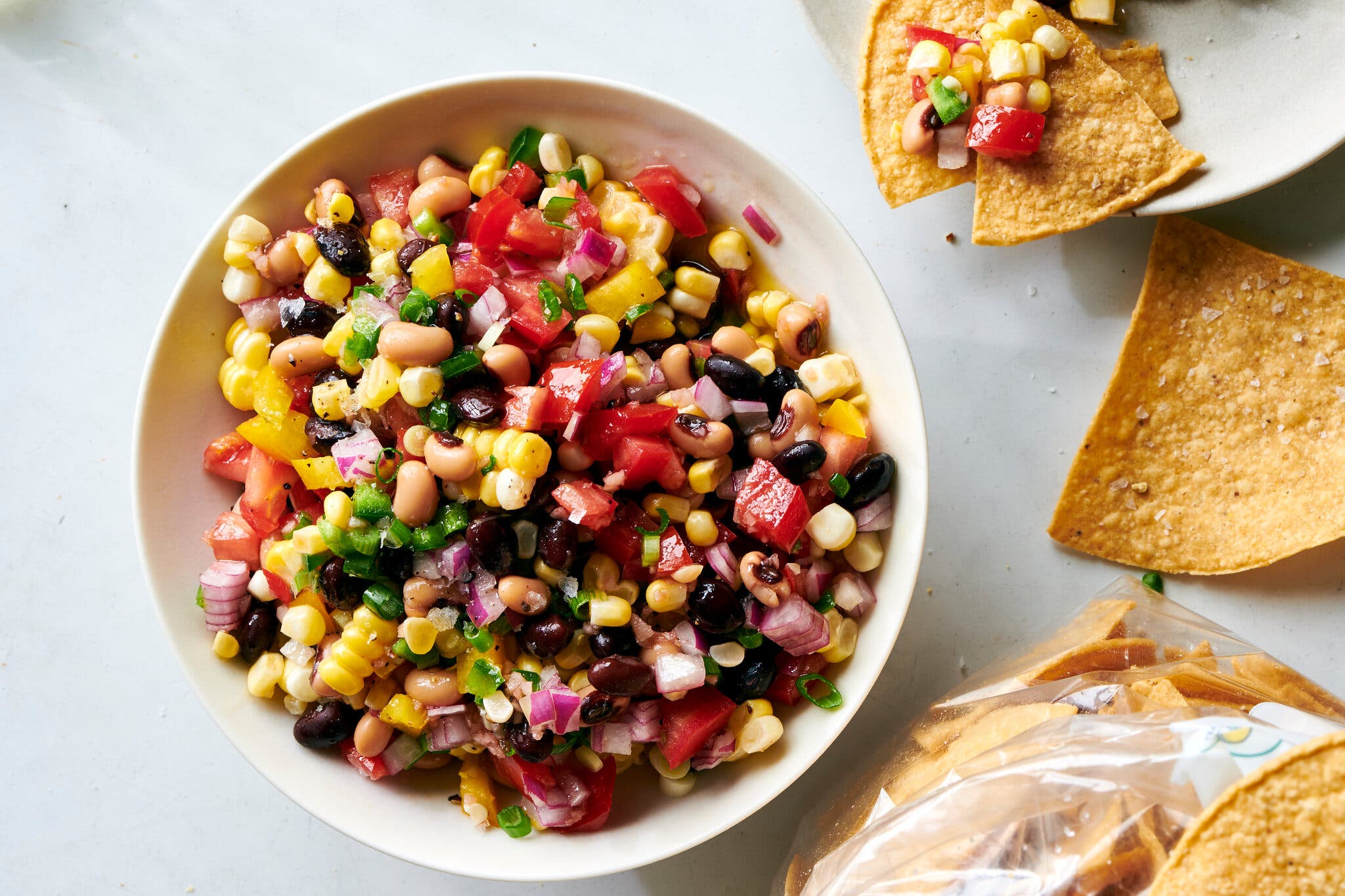 An overhead image of a white bowl filled with chopped red tomatoes and red onion, corn kernels, black beans and black-eyed peas, jalapeño and sliced scallions. Corn tortilla chips are on the side. 