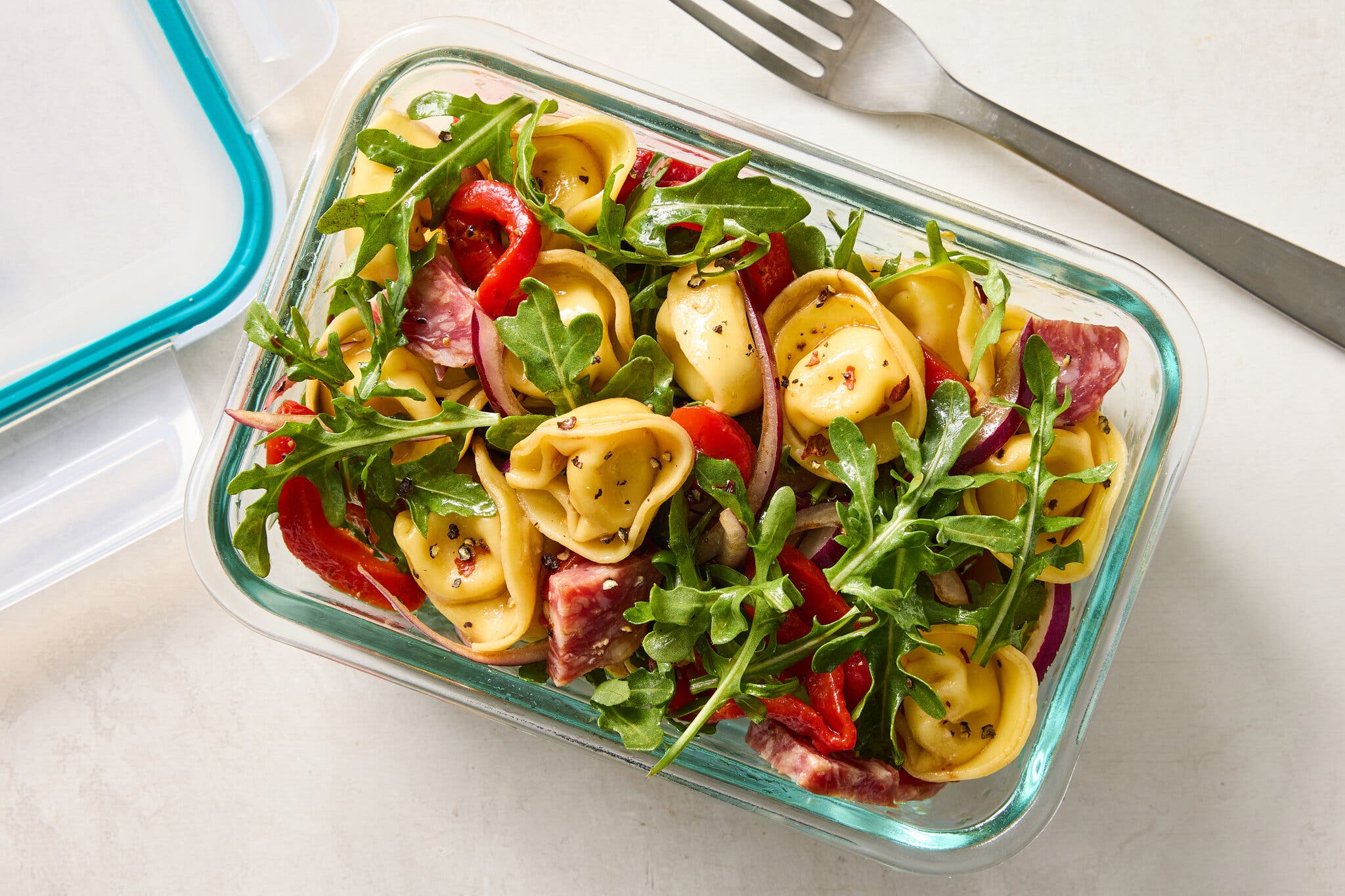An overhead image of a rectangular clear glass container filled with round tortellini pasta, red peppers, salami chunks and arugula leaves.