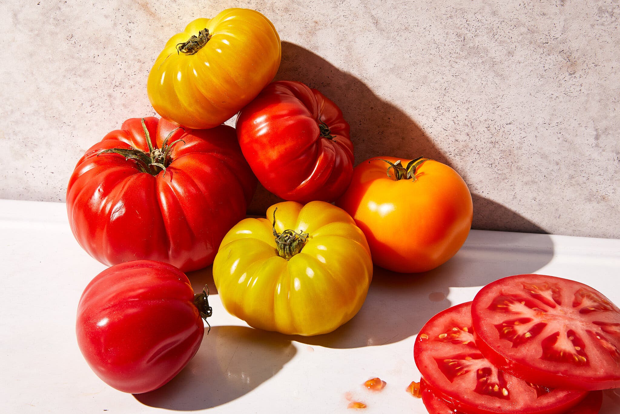A small pyramid of heirloom tomatoes is piled against a wall. Two slices sit off to the bottom corner.
